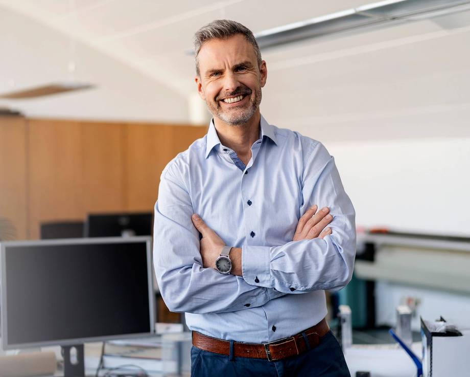 Arbeitserprobung Ein Mann steht in einem Büro mit verschränkten Armen und lächelt.