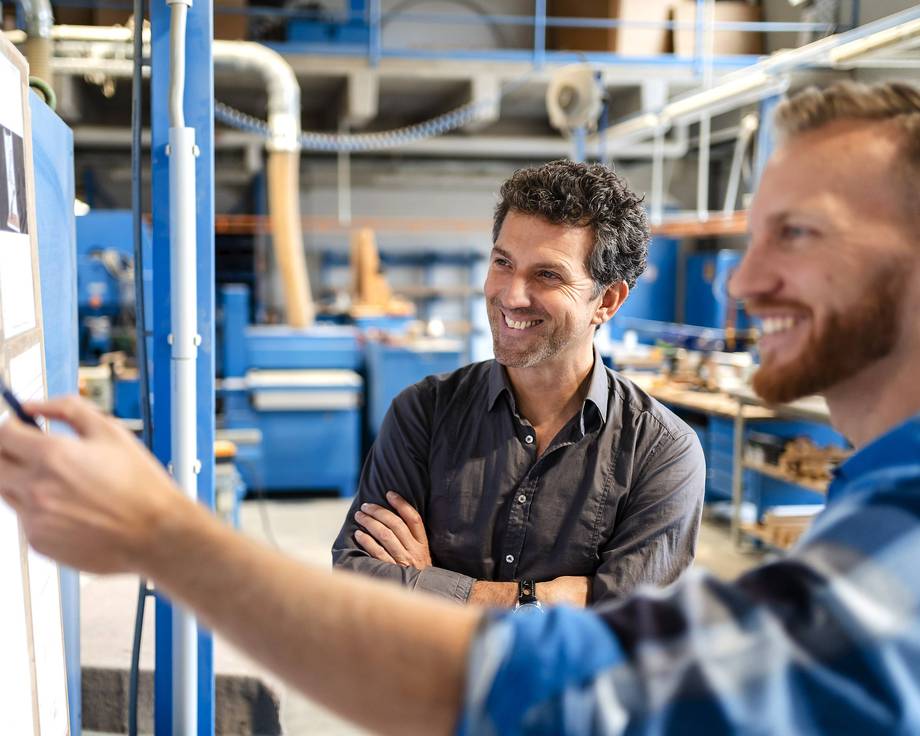 Reha-Vorbereitungslehrgang (RVL) Zwei Männer stehen in einer industriellen Umgebung und schauen auf ein Whiteboard.