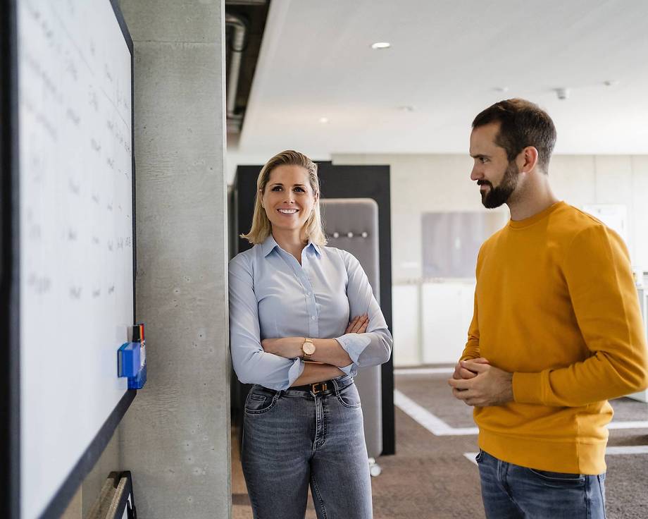 Integration Plus Zwei Personen stehen vor einer Whiteboard-Wand in einem Büro.